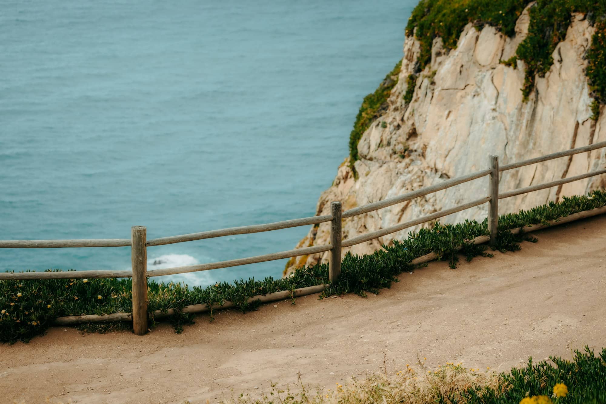 Wooden fence, dirt path, and cliff overlooking the ocean.