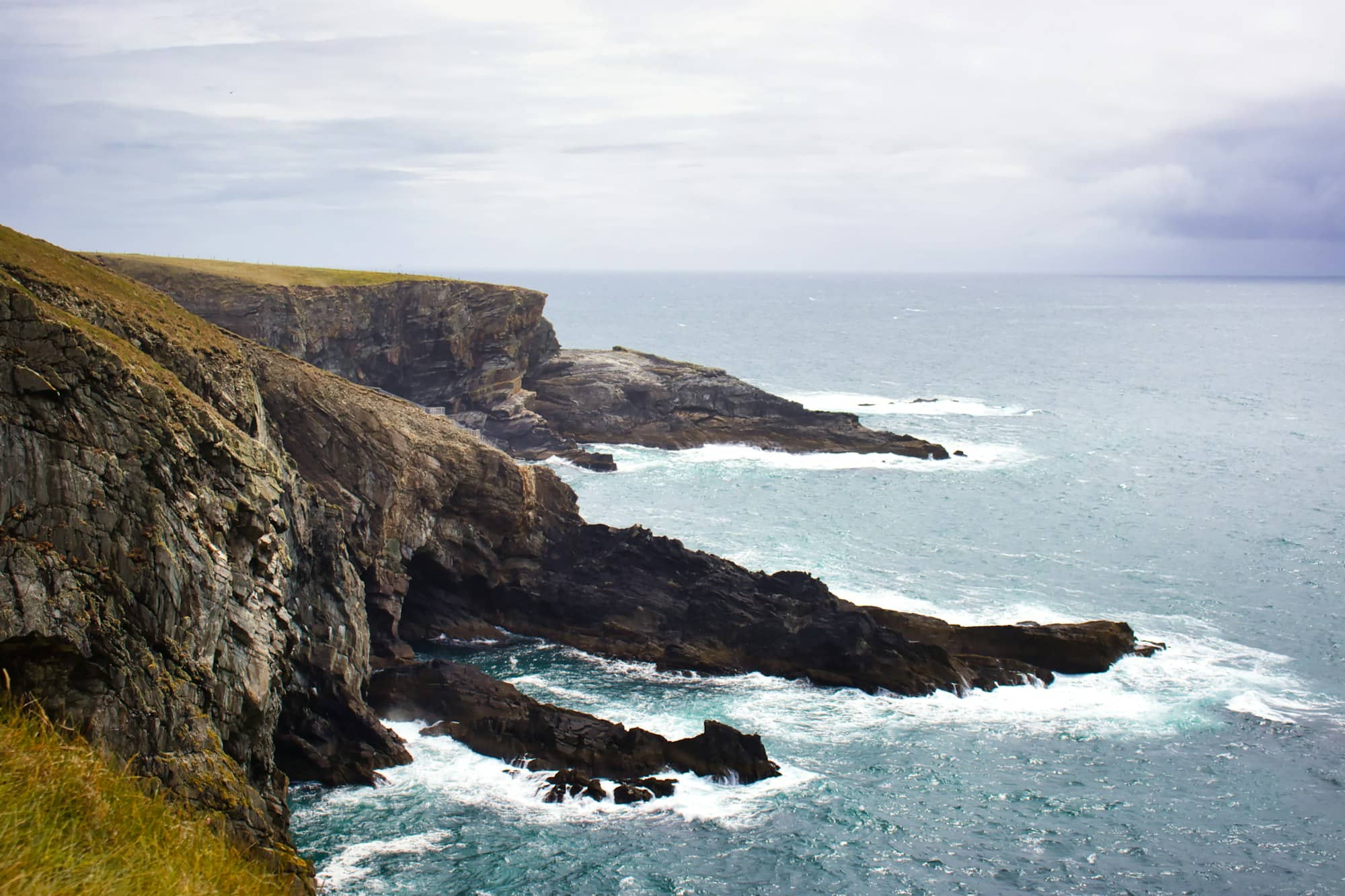 Rugged cliffs meet the churning ocean under a cloudy sky.