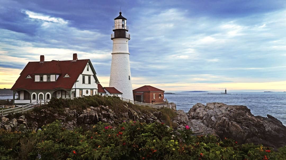 Lighthouse stands on rocky coast with cloudy sky.